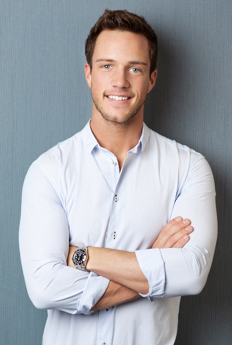 A young man with short brown hair wearing a light blue shirt and a watch, smiling with arms crossed.