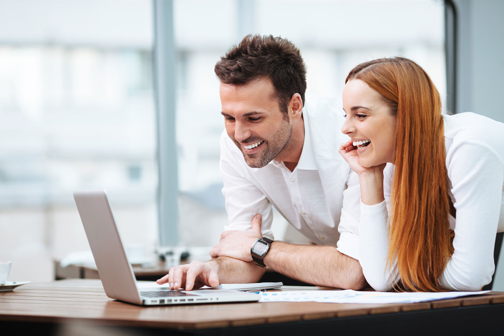 Man and woman smiling while looking at a laptop screen in an office setting.