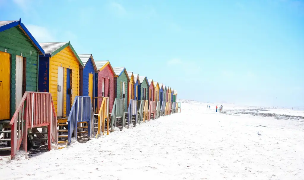 Row of colorful beach huts with wooden steps on a sandy beach under a clear blue sky.